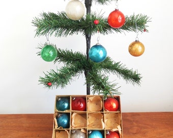 Pode incluir: Uma pequena árvore de Natal artificial decorada com ornamentos de vidro vintage em tons de azul, verde, vermelho e dourado. Os ornamentos estão dispostos em uma caixa com papel de seda.