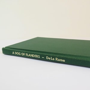 May include: A hardcover book titled "A Dog of Flanders" by De La Rame. The book has a dark green cover with gold lettering. The book is lying on a white surface.