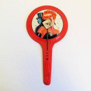 May include: A red metal toy with a round face featuring a woman with red hair and a man in a top hat. The woman's face is partially covered by a metal flap. The toy has a long red handle.