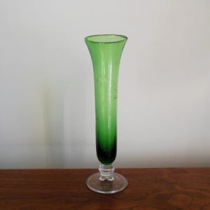 May include: A tall, green glass vase with a clear glass base. The vase has a flared opening and a slender, cylindrical body. The vase is decorated with a subtle etched design.