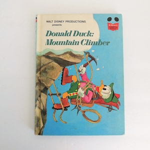 May include: A vintage children's book cover featuring Donald Duck as a mountain climber. The cover is blue with a picture of Donald Duck hanging from a rope on a cliff. The title of the book is "Donald Duck: Mountain Climber".