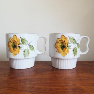 May include: Two white ceramic mugs with a floral design. The mugs have a stacked design with a wide base and a smaller top section. The flowers are yellow with orange centers and green leaves. The mugs have a black rim and a black handle.