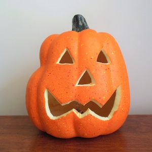 May include: Orange ceramic pumpkin with a carved jack-o'-lantern face. The pumpkin has a speckled orange paint finish and a green stem.