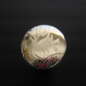 May include: A round, silver-toned object with an etched design of a bird with outstretched wings, surrounded by foliage. The object has a reflective, slightly aged appearance, set against a dark background.