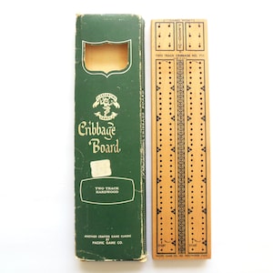 May include: A two-track wooden cribbage board with black printed markings and a matching green cardboard box. The box features the text "Pleasantime Games Cribbage Board" and "Two Track Hardwood," while the board includes "Games Won" and "Two Track Cribbage No. 711" labels.