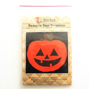 May include: A package containing a pumpkin door decoration applique pattern. The package features a picture of a bright orange pumpkin with a black jack-o-lantern face. The text on the package reads "Patch Press Pumpkin Door Decoration".