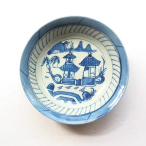 May include: A round, blue and white ceramic bowl with a detailed Asian-inspired landscape design. The bowl features a light blue rim with a textured pattern, and the interior showcases a blue and white illustration of pagodas, trees, and a bridge.
