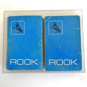 May include: Two blue decks of Rook playing cards in a clear plastic case. The cards have a black crow illustration and the word "ROOK" printed in white.