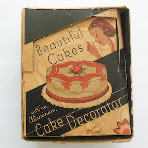 May include: Vintage cake decorator box with the words "Beautiful Cakes" and "Cake Decorator" printed on the front. The box features an illustration of a decorated cake with red accents and a woman admiring a cake.