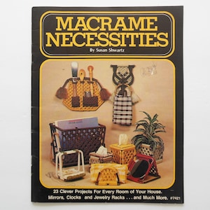 May include: A book cover with the title "Macrame Necessities" by Susan Shwartz. The cover features a variety of macrame projects, including a wall hanging, a plant hanger, a tissue box cover, and a mirror. The text on the cover reads "23 Clever Projects For Every Room of Your House. Mirrors, Clocks and Jewelry Racks... and Much More. #7421"