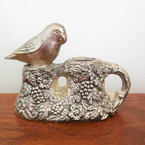 May include: Silver candle holder with a bird perched on top. The base is decorated with grape vines and leaves.