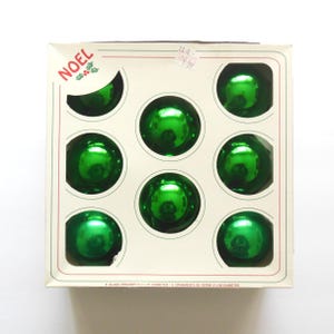 May include: A box of eight shiny green glass ball ornaments, each measuring 2.5 inches in diameter. The white cardboard packaging features the text NOEL with a holly graphic in the corner and a clear window displaying the round ornaments.