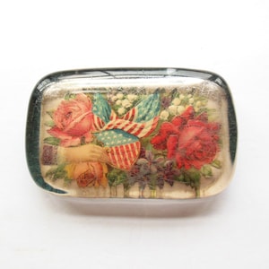 May include: A rectangular glass paperweight featuring a floral design with red and pink roses, white flowers, and American flags. The design includes a hand holding a shield with stars and stripes.