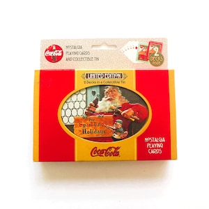 May include: A limited edition Coca-Cola nostalgia playing cards set in a collectible tin. The packaging features a red and yellow design with an illustration of Santa Claus holding a bottle and a sign that reads For Sparkling Holidays.
