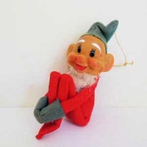 May include: A vintage Christmas elf doll with a plastic face, blue eyes, and a red outfit. The elf has a green hat and mittens, and a white collar. The doll is in a sitting position.