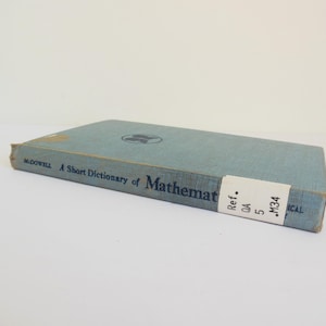 May include: A light blue hardcover book titled A Short Dictionary of Mathematics by McDowell. The spine features a white library call number label with the text Ref. QA 5 .M34. A small circular logo is visible on the front cover.