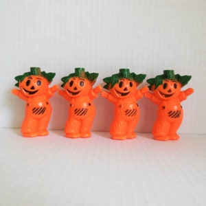 May include: Four orange plastic jack-o'-lantern figurines with green leaf stems and black painted faces. The figurines are standing in a row on a white surface.
