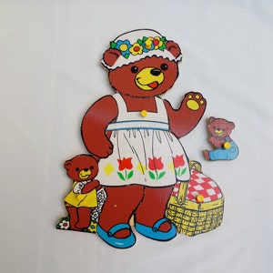 May include: A wooden toy set featuring a brown bear wearing a white dress with red flowers, a flower crown, and blue shoes. The bear is holding a basket with a red and white checkered lid. There is a smaller brown bear wearing a blue shirt and blue pants.