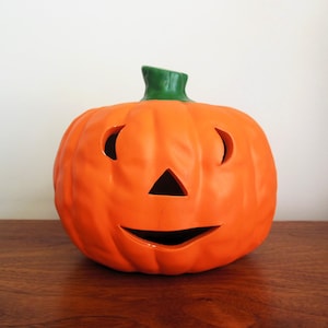 May include: Orange ceramic jack-o'-lantern with a green stem. The pumpkin has cut-out eyes, a triangular nose, and a smiling mouth. A festive Halloween decoration.