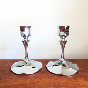 May include: Two polished silver candle holders. Each has a faceted cup, a slender stem, and a wide, geometric base. The candle holders are displayed on a wooden surface, reflecting the light.