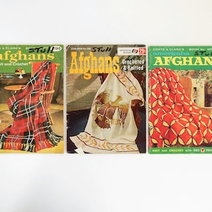 May include: Three vintage crochet afghan pattern books with colorful covers featuring different designs. The covers show a red and black plaid afghan, a white afghan with an eagle design, and a red and yellow geometric afghan. The books are titled "Afghans" and feature the "Red Heart" yarn brand.