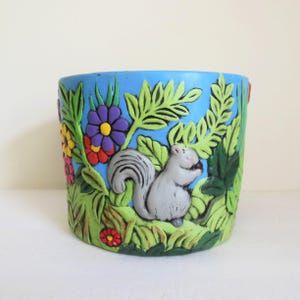 May include: A blue ceramic planter with a 3D design of a squirrel, flowers, and leaves. The squirrel is gray and holding a nut. The flowers are red, yellow, purple, and orange.