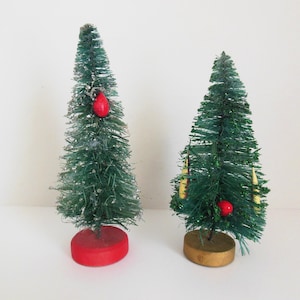 May include: Two miniature Christmas trees. The trees are green with a flocked appearance and are decorated with red ornaments and gold-colored candle-shaped ornaments. One tree has a red base, and the other has a gold base.