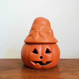 May include: Orange ceramic pumpkin-shaped container with a lid. The pumpkin has carved triangular eyes, a round nose, and a smiling mouth. The lid is shaped like a hat with a flower design. The container is on a wooden surface.