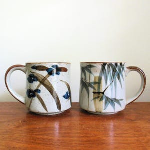 May include: Two ceramic mugs with brown handles and rims. One mug features blue floral designs and brown brushstrokes on a speckled white background. The other mug has a bamboo design in shades of green and brown.