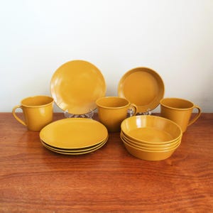 May include: A mustard yellow dinnerware set, including plates, bowls, and mugs. The set features a classic design with a glossy finish. The plates are stacked, and the bowls are nested. The mugs have handles. The set is arranged on a wooden surface.