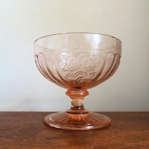 May include: A vintage pink glass dessert bowl with a floral design. The bowl has a rounded shape with a pedestal base and a detailed floral pattern etched into the glass. The bowl is made of pink glass.