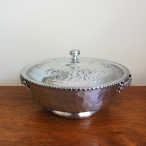 May include: A round, silver-toned metal serving bowl with a matching lid. The bowl features a hammered texture on the exterior and two side handles. The lid is decorated with an embossed grape and leaf motif and a central knob handle.