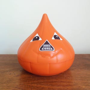 May include: Orange Hershey's Kisses candy container shaped like a Hershey's Kiss with a smiling face and eyes. The container has a label that says "Hershey's Milk Chocolate Kisses". The container is sitting on a wooden surface.