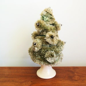 May include: A small, green, flocked Christmas tree with white lights. The tree is on a white ceramic base.