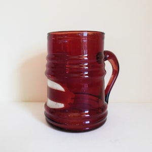 May include: A vintage, ruby-red glass mug with a handle. The mug features horizontal ridges around the body and a white, abstract design on the front. The glass has a slight transparency, and the mug is likely used for beverages.