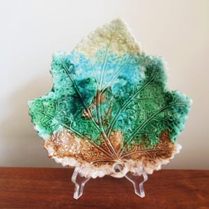 May include: A ceramic leaf-shaped dish with a textured surface. The dish features a color gradient from brown to green to blue, with visible leaf vein details. It is displayed on a clear acrylic stand, showcasing its decorative design.