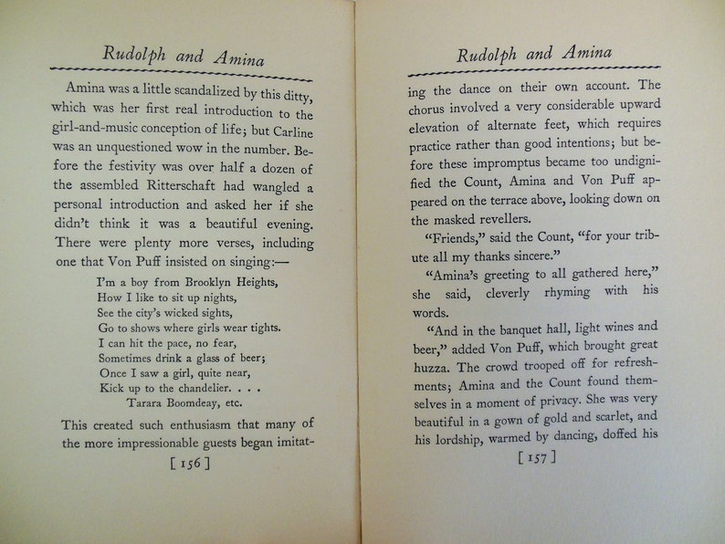 May include: A black and white book with the title "Rudolph and Amina" on the cover. The book is open to a page with text about a character named Amina and a song she sings. The page numbers are 156 and 157.