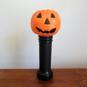 May include: Orange plastic jack-o'-lantern with a black face and a black handle. The jack-o'-lantern is on a wooden surface.