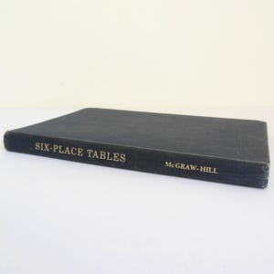 May include: A dark blue hardcover book titled Six-Place Tables with the publisher name McGraw-Hill printed in gold lettering on the spine. The book is positioned horizontally, showcasing the front cover and the spine against a plain background.