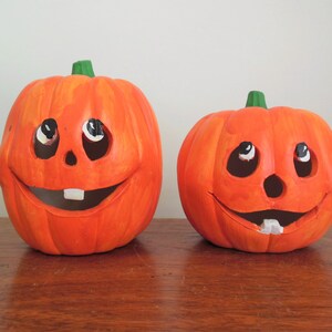 May include: Two ceramic Halloween jack-o'-lanterns with painted faces. The larger pumpkin has a wide smile with a single tooth, while the smaller pumpkin has a smaller smile with a single tooth. Both pumpkins have black eyes and a black nose.