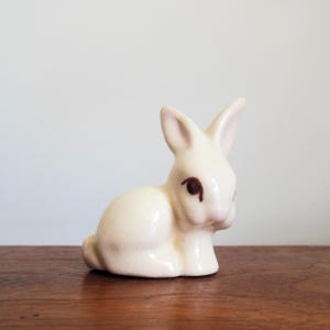 May include: A white ceramic rabbit figurine with brown painted eyes. The rabbit is sitting with its head down and its ears are perked up.