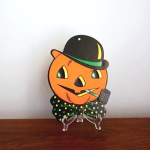 May include: A vintage Halloween decoration of a pumpkin with a black bowler hat, green bow tie with yellow polka dots, and a pipe. The pumpkin has a smiling face with orange skin, black eyes, and a black nose.