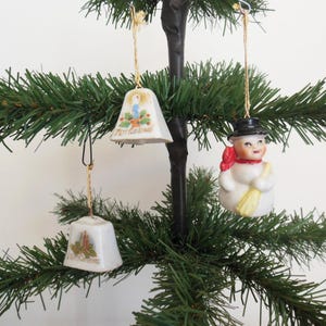 May include: Three vintage Christmas ornaments hanging on a small artificial tree. The ornaments include two white bell-shaped decorations with "Merry Christmas" text and a snowman figure with a black hat and red scarf.