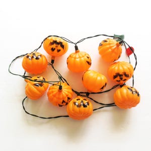 May include: A string of orange pumpkin-shaped lights with black jack-o'-lantern faces. The lights are strung together with green wire.