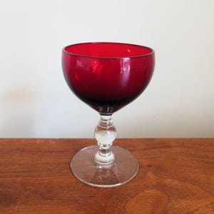 May include: A deep red wine glass with a clear glass stem and base. The stem has a decorative knob.