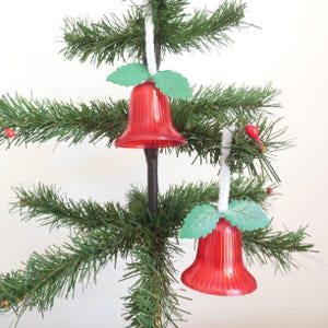 May include: Two red glass bell ornaments with green leaf accents hanging on a green artificial Christmas tree.