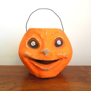 May include: Vintage orange paper mache Halloween jack-o'-lantern with a wire handle. The jack-o'-lantern has a smiling face with white eyes and a black nose.