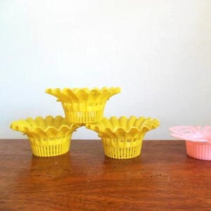 May include: Three bright yellow plastic baskets with scalloped edges are stacked on a wooden surface. The baskets have a flower-like shape and a perforated design. A pink basket is also visible.