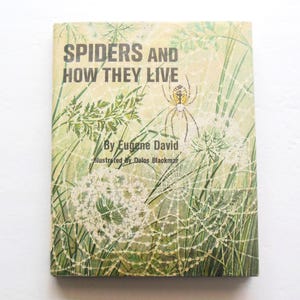 May include: A vintage book cover with a green and yellow spider web design. The title is "Spiders and How They Live" by Eugene David, illustrated by Delos Blackmar.