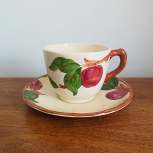 May include: A white teacup and saucer with a brown rim and a floral design featuring red apples and green leaves.